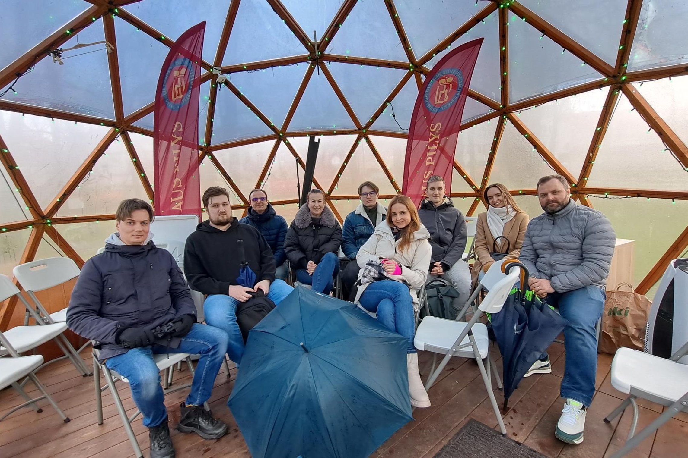 Grupė suaugusiųjų sėdi kupolo formos paviljone Vilniaus pilių teritorijoje, klausosi ekskursijos, laikydami skėčius ir vilkėdami šiltus drabužius