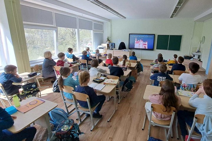 Klasėje mokiniai sėdi prie suolų ir žiūri į priekyje esantį ekraną, kuriame rodoma pamokos medžiaga apie pilis; šalia ekrano – lenta, mokytojo stalas, o palei sienas – spintelės su mokymo priemonėmis ir dideli langai.