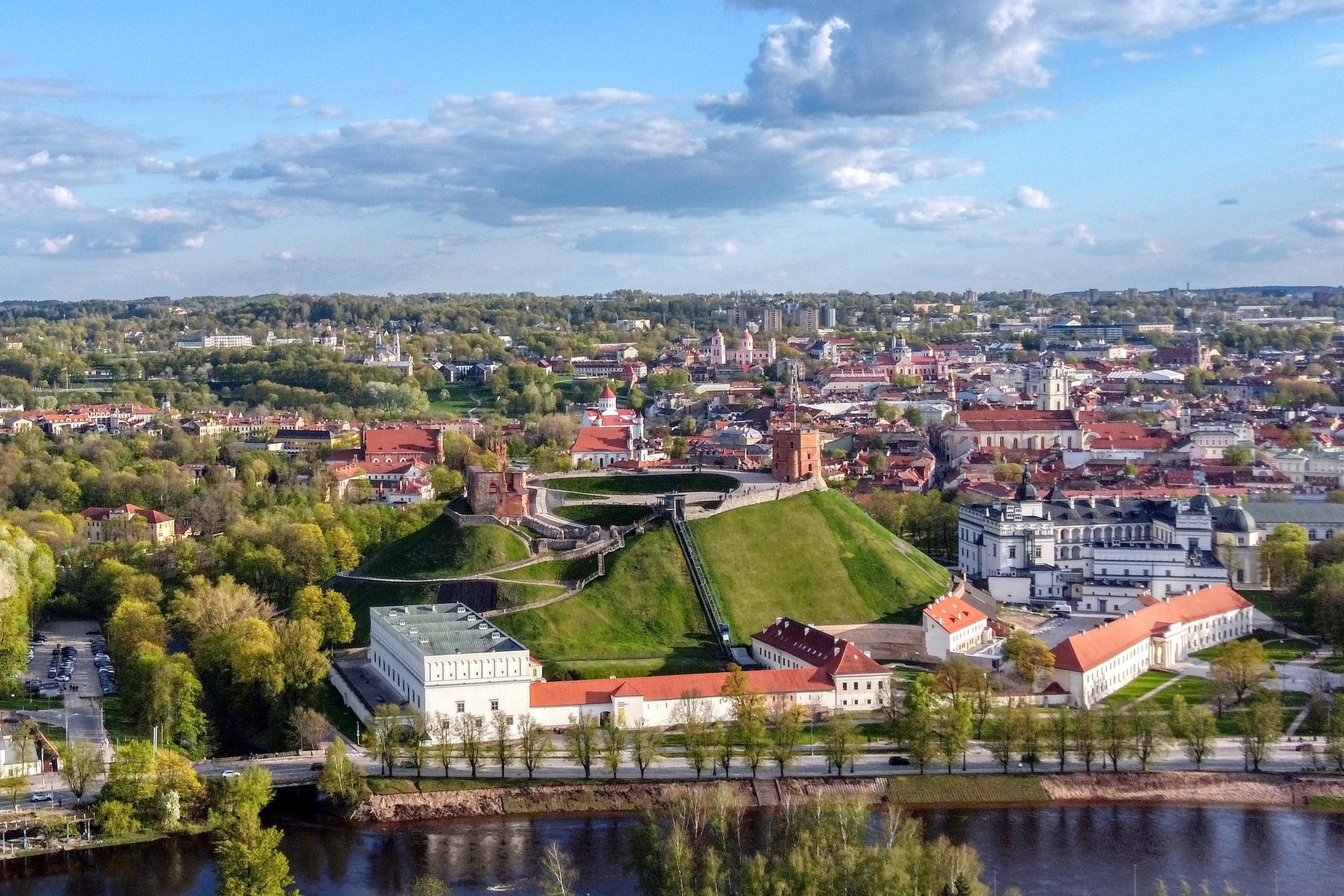 Plati Vilniaus pilių teritorijos panorama iš oro: centre iškyla Gedimino kalnas su Aukštutinės pilies bokštu ir takais, papėdėje matyti Žemutinės pilies pastatai ir kiemai, apačioje teka Neris, o aplink driekiasi žalias parkas ir Vilniaus senamiesčio stog