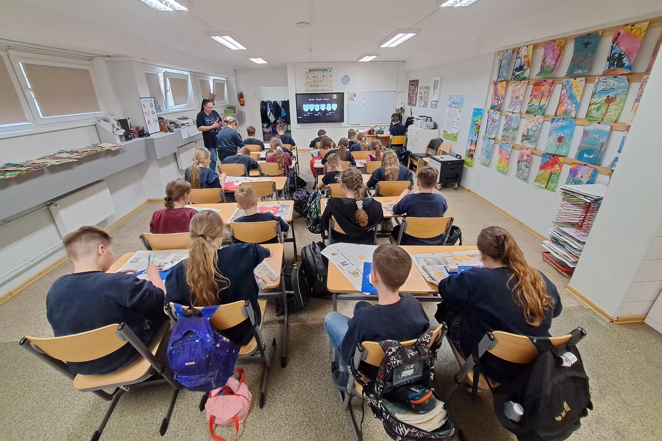 Mokiniai klasėje dalyvauja edukaciniame užsiėmime, sėdi prie suolų ir klausosi pristatymo ekrane, ant sienų eksponuojami spalvingi vaikų piešiniai.