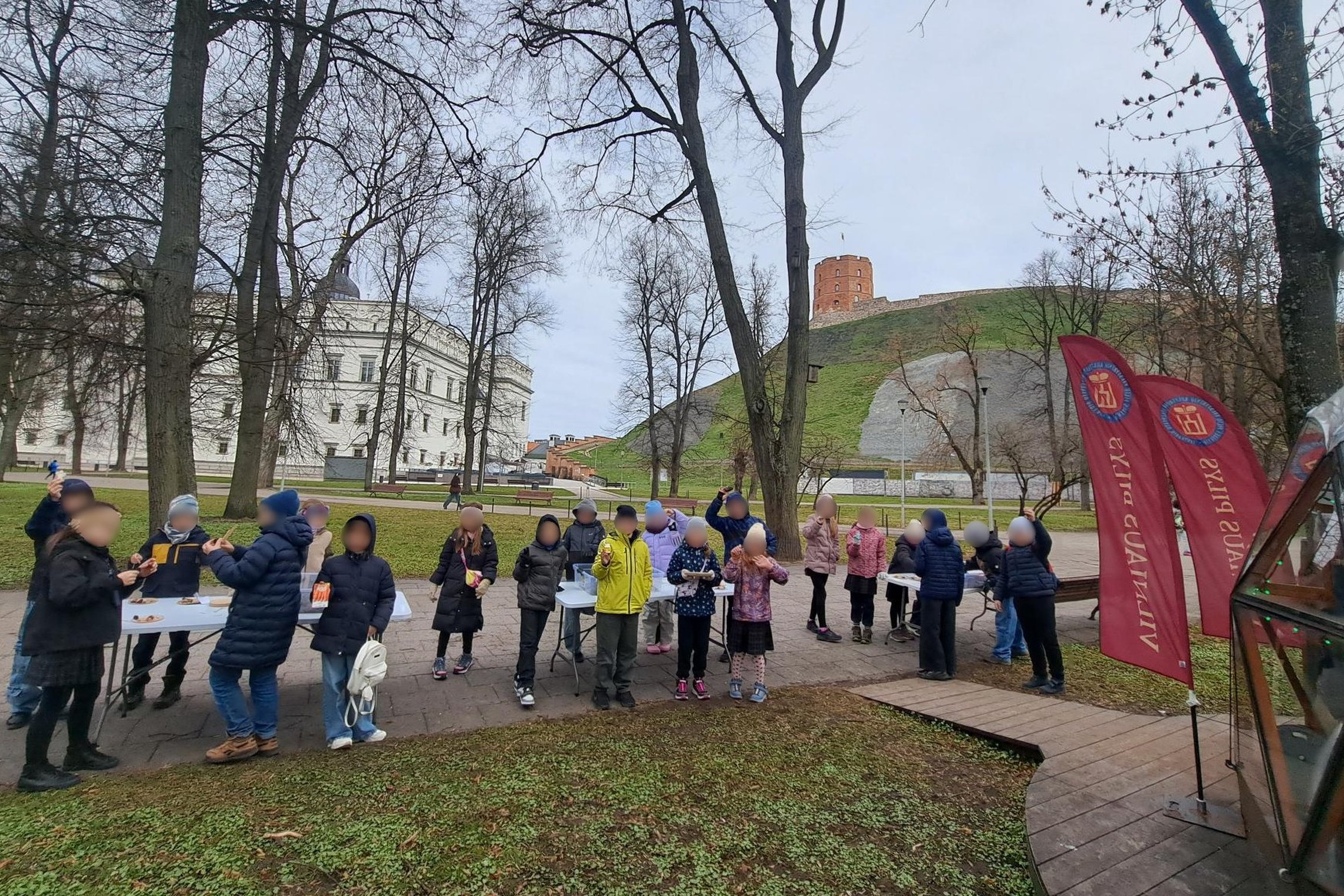 Nuotraukoje vaikai prie Vilniaus pilių kupolo. Jie stovi aplink stalus, ant stalų dėžės su žemės sluoksniu ir radiniais.