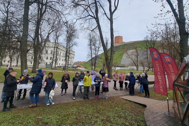Nuotraukoje vaikai prie Vilniaus pilių kupolo. Jie stovi aplink stalus, ant stalų dėžės su žemės sluoksniu ir radiniais.