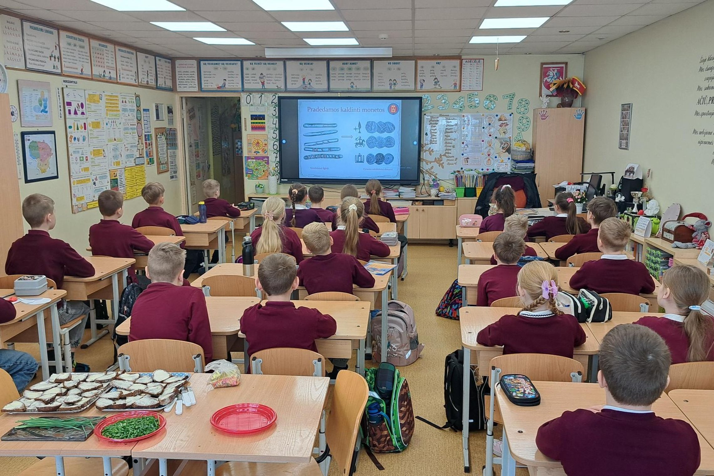 Kauno Kazio Griniaus progimnazijos klasėje mokiniai sėdi suoluose ir stebi interaktyviame ekrane rodomą edukacinę prezentaciją apie senovines monetas. Klasėje matyti mokymo priemonės, mokinių kuprinės ir ant stalo padėtos vaišės.