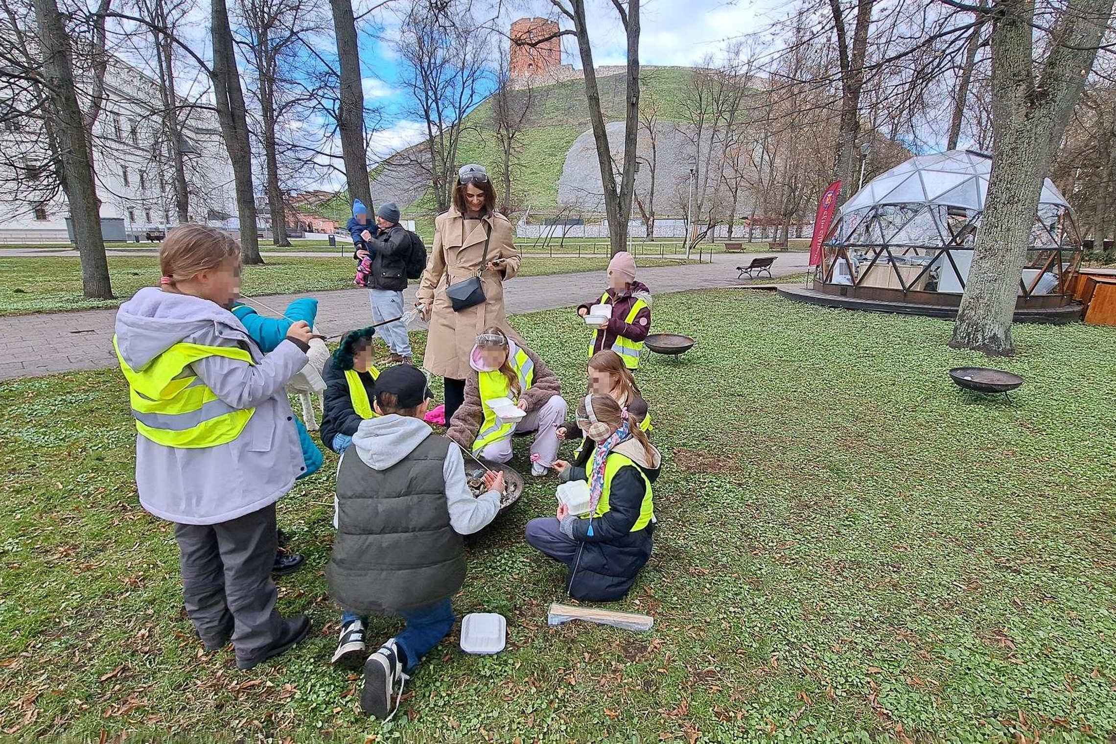 Vaikai su atšvaitinėmis liemenėmis susibūrę parke prie nedidelės laužavietės kepa zefyrus, šalia stovi edukatorė; fone matyti Gedimino pilies kalnas ir kupolo formos stiklinė pavėsinė.