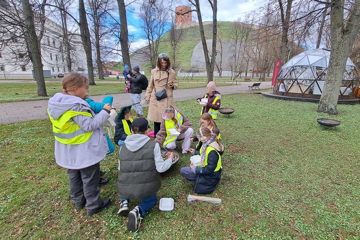 Vaikai su atšvaitinėmis liemenėmis susibūrę parke prie nedidelės laužavietės kepa zefyrus, šalia stovi edukatorė; fone matyti Gedimino pilies kalnas ir kupolo formos stiklinė pavėsinė.