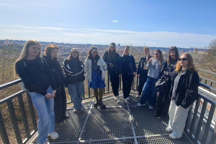 Studentų grupė stovi ant apžvalgos aikštelės su metalinėmis grotelėmis, fone atsiveria Vilniaus miesto panorama su senamiesčiu, pastatais ir mėlynu dangumi.