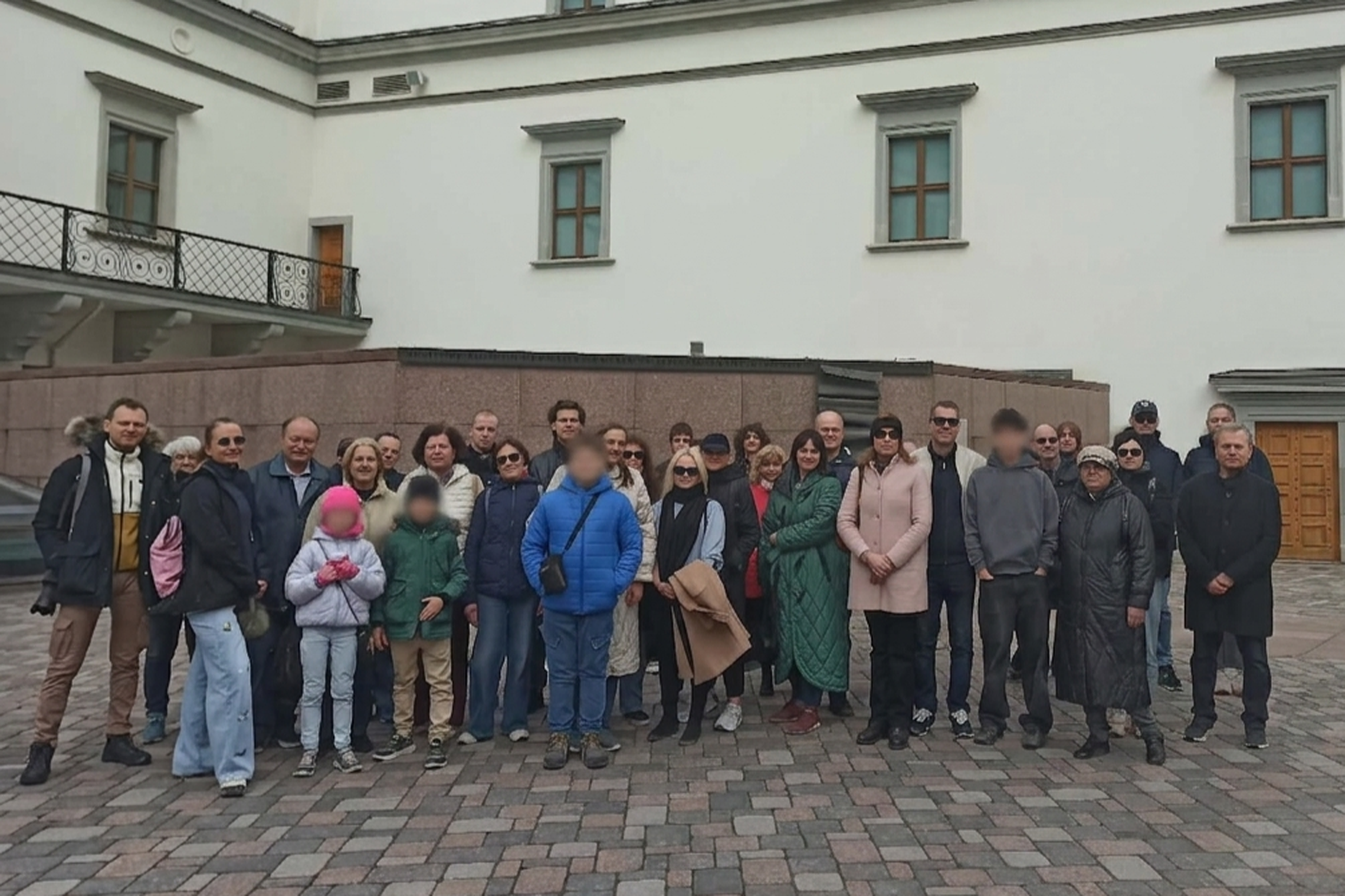 Grupinė ekskursijos dalyvių nuotrauka Vilniaus pilių teritorijoje prie pastato – suaugusieji ir vaikai stovi kieme priešais šviesų fasadą.