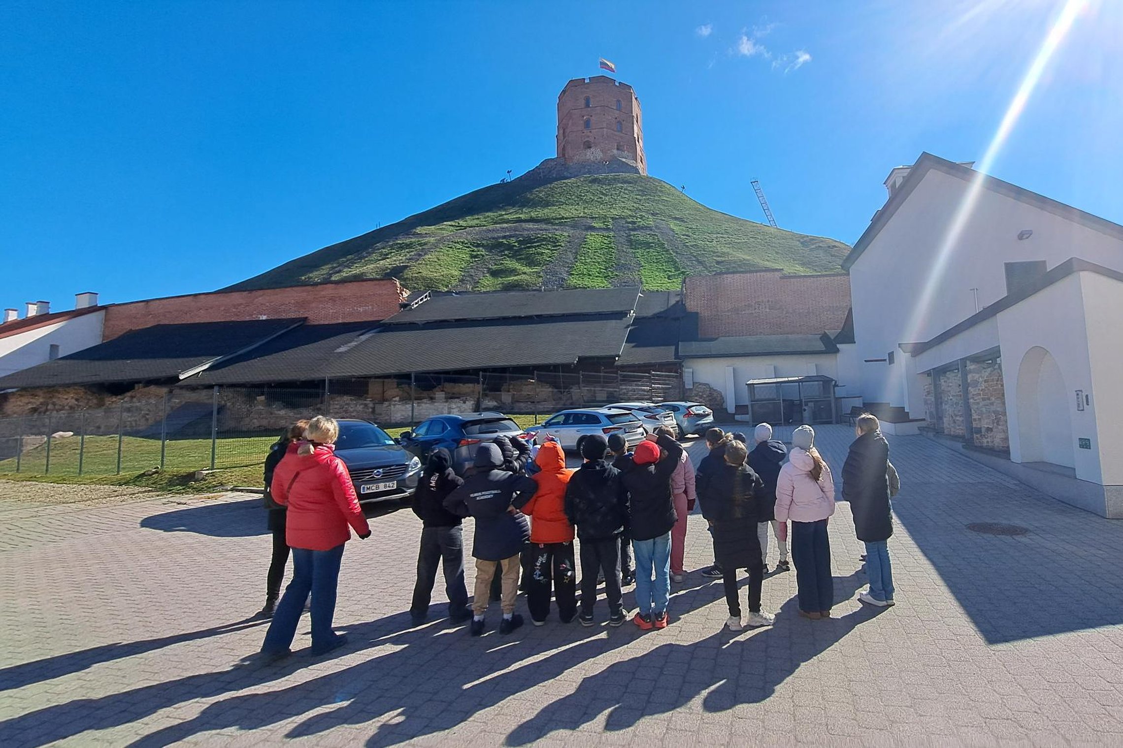 Grupė pradinių klasių mokinių su mokytojais stovi Vilniaus pilių teritorijoje ir žvelgia į Gedimino kalną su bokštu saulėtą dieną.