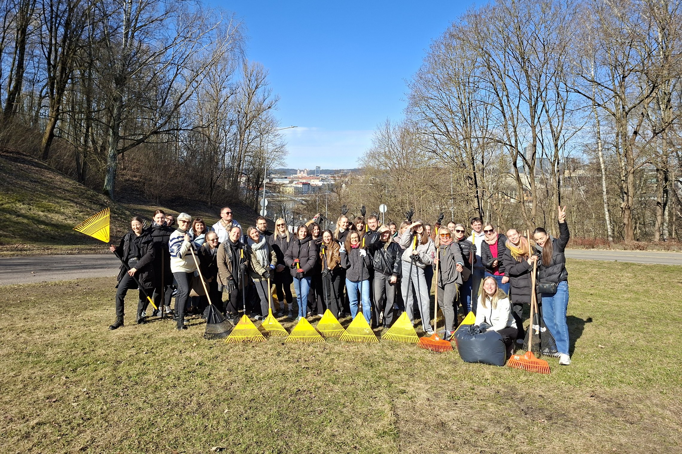 Didelė savanorių grupė su grėbliais ir šiukšlių maišais stovi Kalnų parke saulėtą pavasario dieną, apsupta dar plikų medžių. Visi šypsosi, mojuoja ir pozuoja bendram nuotraukos kadrui po talkos.