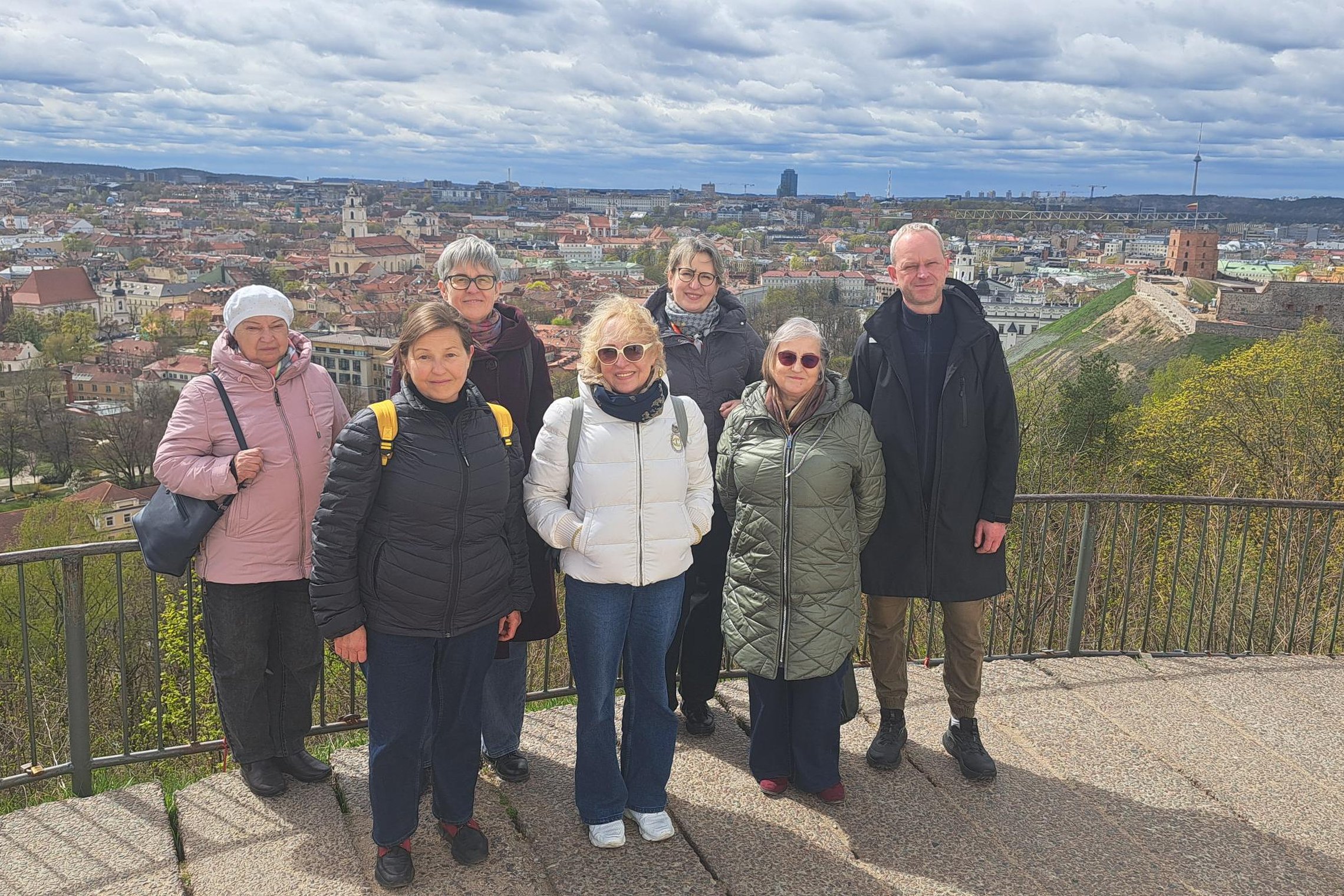 Ekskursijos dalyviai stovi apžvalgos aikštelėje Vilniaus pilių teritorijoje, fone atsiveria Vilniaus senamiesčio panorama ir Gedimino pilies bokštas.