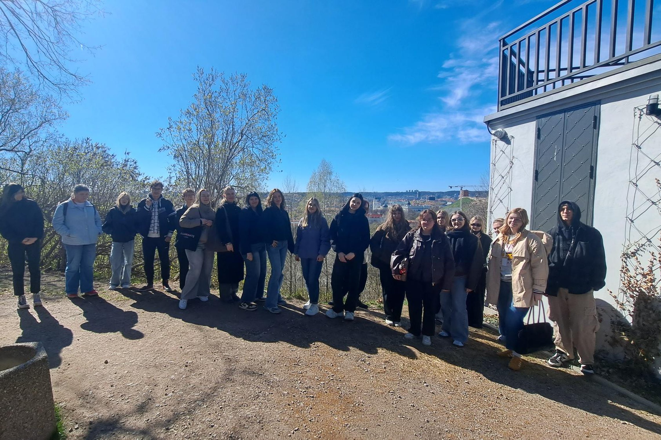Vilniaus pilių teritorijoje saulėtą dieną stovi Vilniaus kolegijos studentų grupė, fone matyti medžiai, miesto panorama ir pastato siena su durimis.