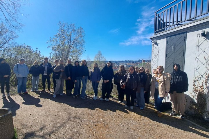 Vilniaus pilių teritorijoje saulėtą dieną stovi Vilniaus kolegijos studentų grupė, fone matyti medžiai, miesto panorama ir pastato siena su durimis.