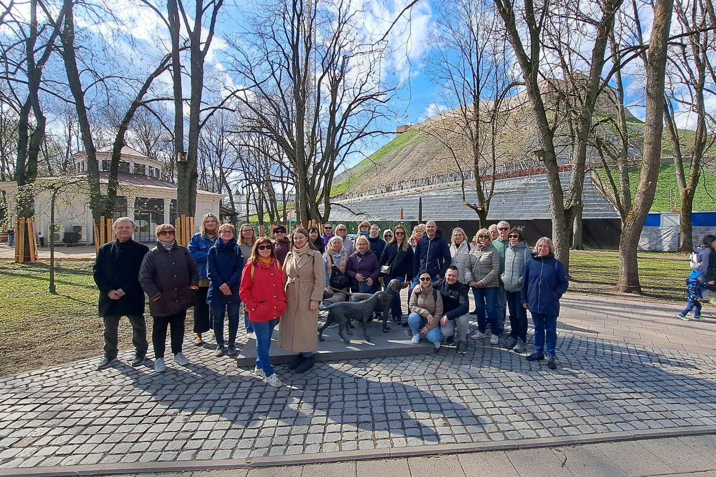 Grupė žmonių stovi lauke prie paminklo su šunimis, už jų matyti medžiai, pastatas ir žaliuojantis kalnas su takais.