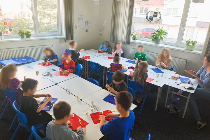 Nuotraukoje šviesi ir jauki klasė. Joje stovi stalai, prie kurių sėdi vaikai. Vaikai dalyvauja edukacijoje.