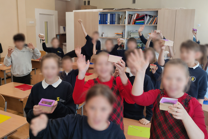 Mokiniai klasėje pakėlę rankas džiaugsmingai rodo savo pačių pagamintus spalvingus muilus edukacijos metu.