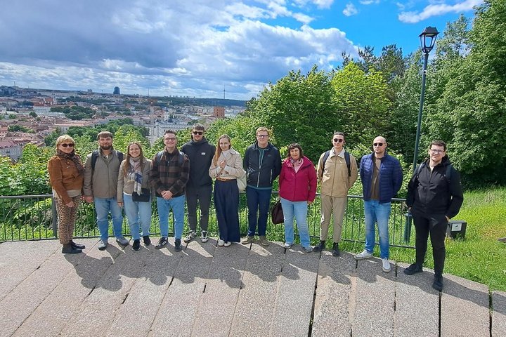 Ekskursijos dalyviai Vilniaus pilių teritorijoje stovi apžvalgos aikštelėje su atsiveriančia Vilniaus senamiesčio panorama. Fone matyti miesto bokštai, raudoni stogai ir žaliuojančios kalvos, o virš jų driekiasi mėlynas dangus su dideliais baltais debesim