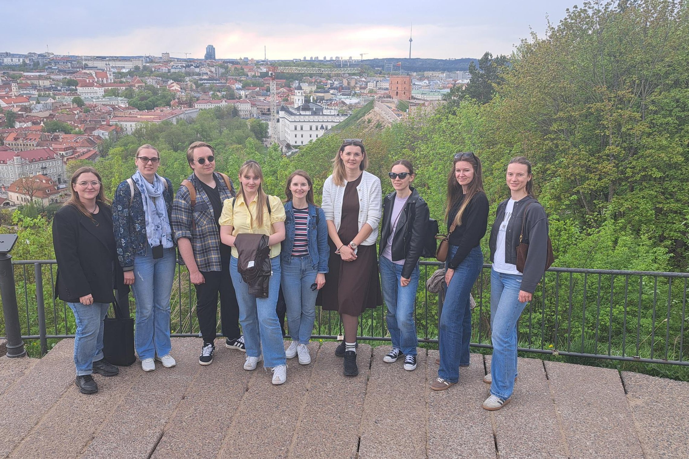 Grupė žmonių stovi apžvalgos aikštelėje Vilniaus pilių teritorijoje, fone matyti senamiesčio panorama su bažnyčių bokštais ir Gedimino pilies kalnu, aplink žaliuojantys medžiai.