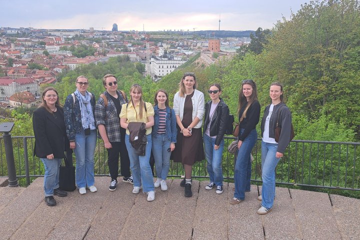 Grupė žmonių stovi apžvalgos aikštelėje Vilniaus pilių teritorijoje, fone matyti senamiesčio panorama su bažnyčių bokštais ir Gedimino pilies kalnu, aplink žaliuojantys medžiai.