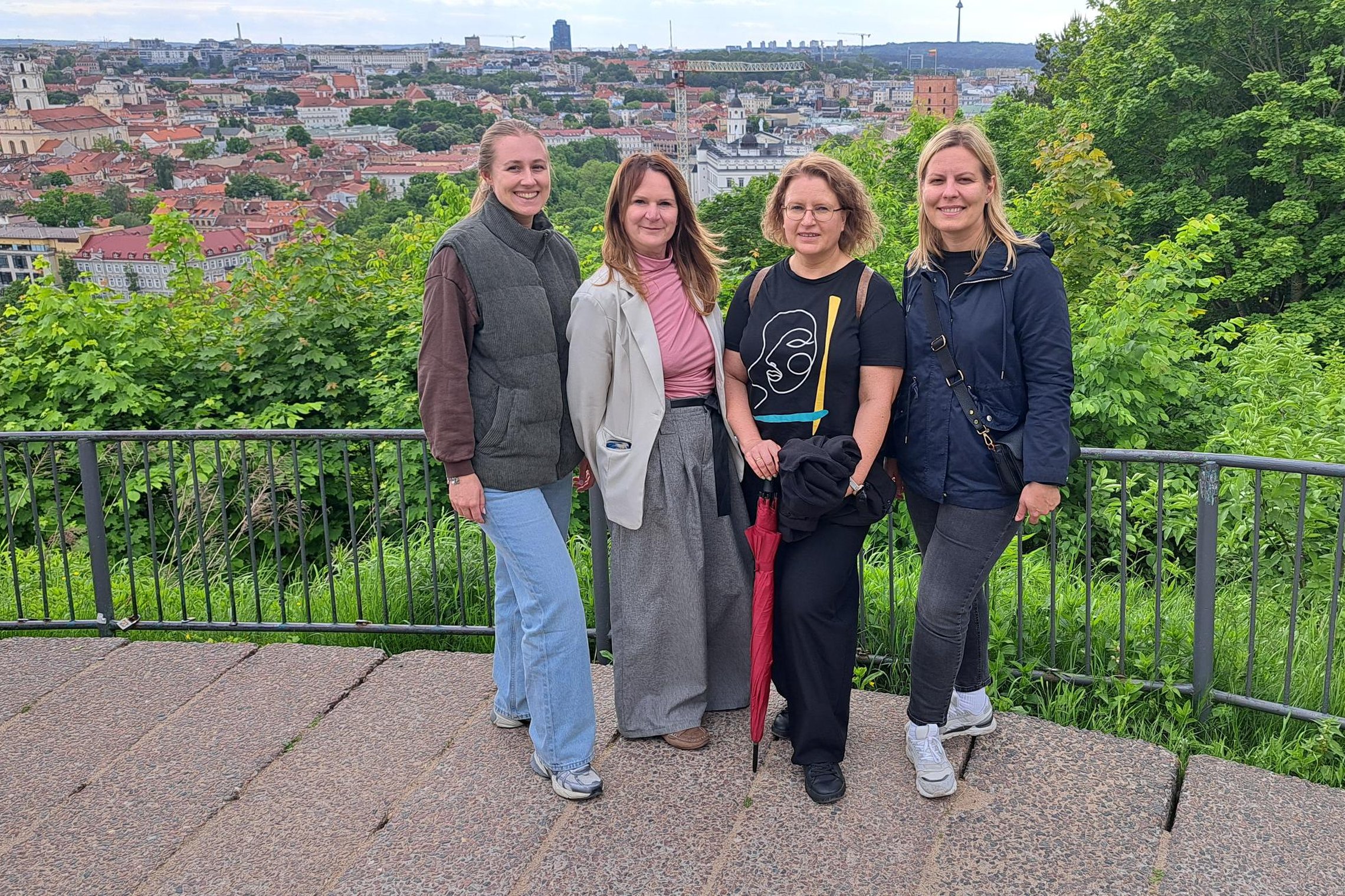 Keturių žmonių grupė stovi Vilniaus pilių teritorijos apžvalgos aikštelėje. Už jų atsiveria Vilniaus senamiesčio panorama su istoriniais pastatais, Gedimino pilies bokštu ir žaliuojančiomis kalvomis.