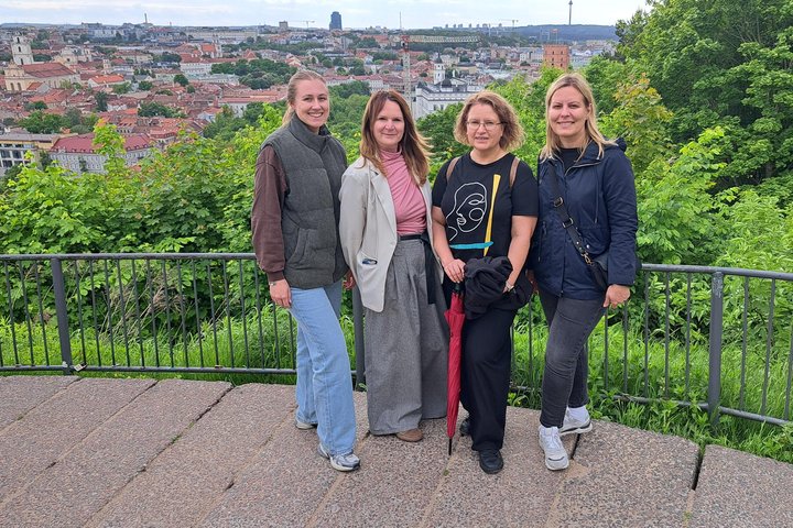 Keturių žmonių grupė stovi Vilniaus pilių teritorijos apžvalgos aikštelėje. Už jų atsiveria Vilniaus senamiesčio panorama su istoriniais pastatais, Gedimino pilies bokštu ir žaliuojančiomis kalvomis.