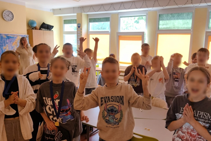 Klasėje stovi būrys vaikų, keliančių rankas ir rodančių gestus, dalis jų su medaliais ant kaklo. Fone matyti mokytoja, žemėlapis, spintelės ir dideli langai su nuleistomis žaliuzėmis.