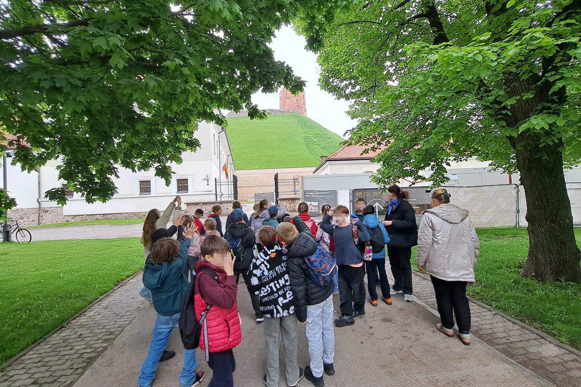 Vaikų grupė dalyvauja edukacijoje Vilniaus pilių teritorijoje. Fone matyti žalias Gedimino kalnas ir Aukštutinės pilies bokštas, o mokiniai klausosi edukatorės pasakojimo po medžiais apsuptame take.