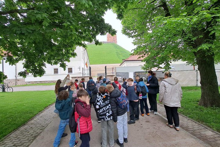 Vaikų grupė dalyvauja edukacijoje Vilniaus pilių teritorijoje. Fone matyti žalias Gedimino kalnas ir Aukštutinės pilies bokštas, o mokiniai klausosi edukatorės pasakojimo po medžiais apsuptame take.