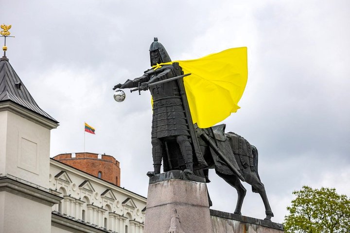 Kunigaikščio Gedimino paminklas Vilniaus pilių teritorijoje, papuoštas ryškiai geltonu apsiaustu. Fone matyti Gedimino pilies bokštas, Lietuvos trispalvė ir debesuotas dangus.