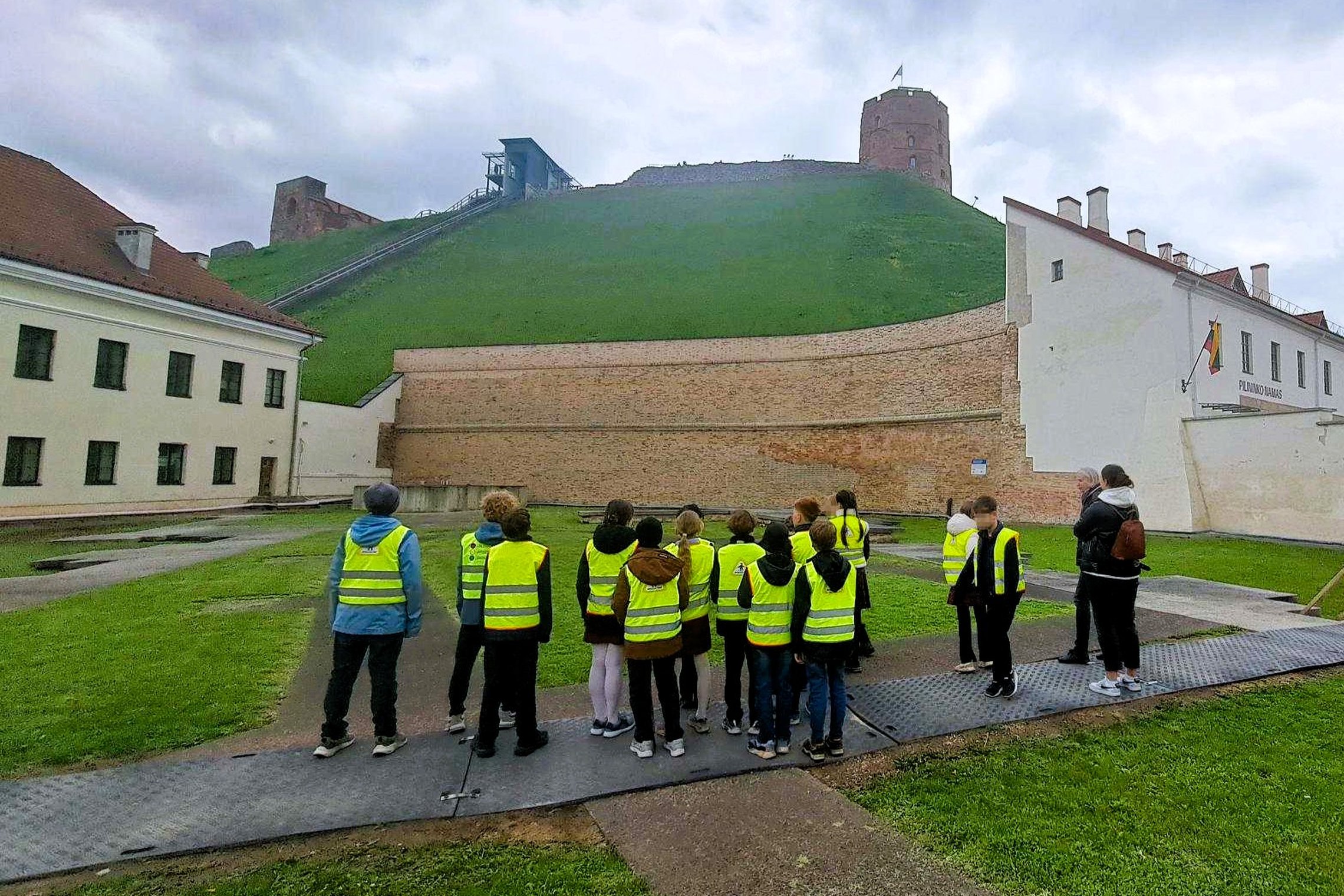 Vaikai su ryškiomis geltonomis liemenėmis stovi Vilniaus pilių teritorija kieme ir klausosi edukacijos prie Gedimino kalno bei Gedimino pilies bokštas. Virš kalno matyti apsiniaukęs dangus, o aplink – istoriniai mūriniai pastatai ir žalios vejos plotai.