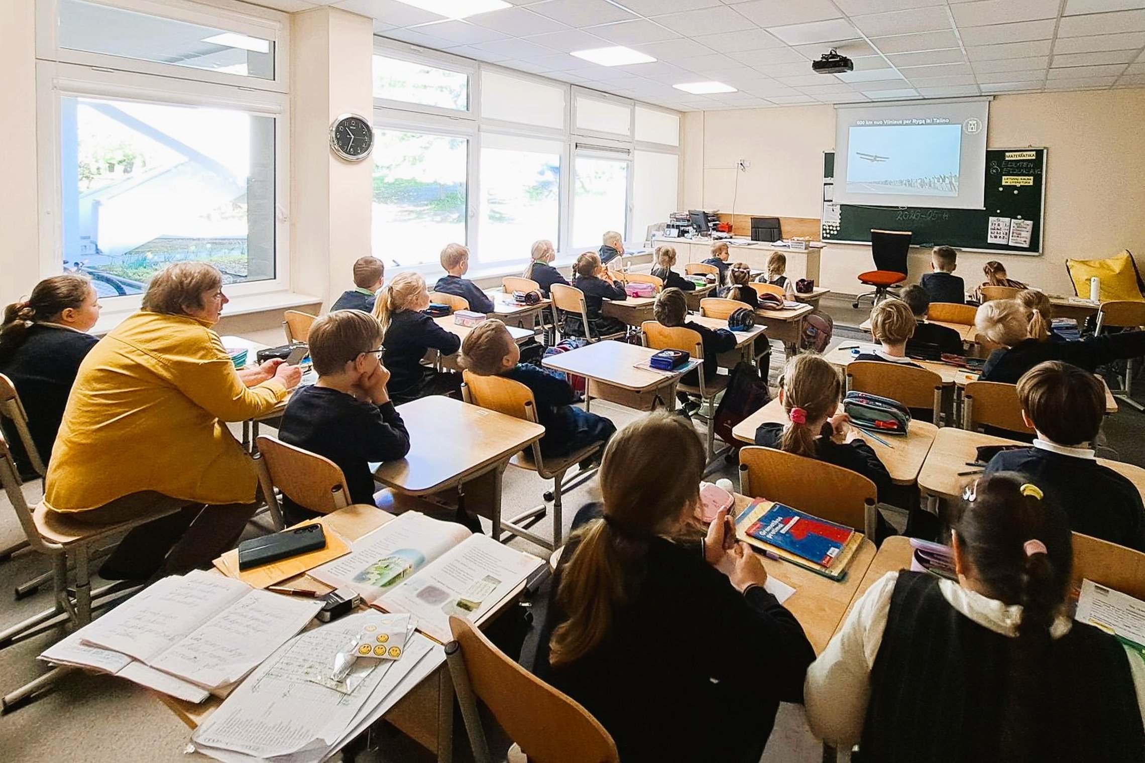 Nuotraukoje mokiniai dalyvauja edukacijoje „Baltijos kelias Vilniaus pilyse“. Jie sėdi suoluose, žiūri į pristatymą ekrane. Ekrane juodai balta Baltijos kelio nuotrauka su žmonių grandine ir virš jų skrendančiu lėktuvu.
