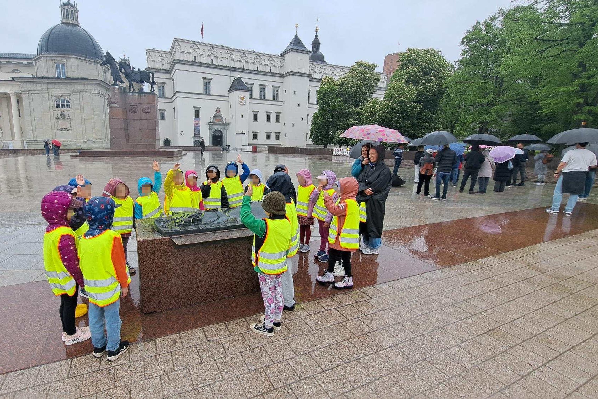 Vaikai su ryškiomis liemenėmis klausosi edukacijos Katedros aikštėje lietingą dieną, šalia Vilniaus pilių maketo.