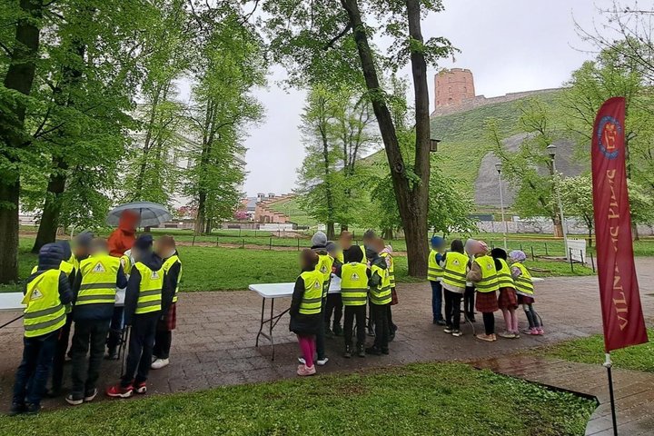 Vaikai su ryškiomis liemenėmis dalyvauja edukacijoje prie Vilniaus pilių kupolo, o fone matyti Gedimino pilies bokštas ir žaliuojantys medžiai.