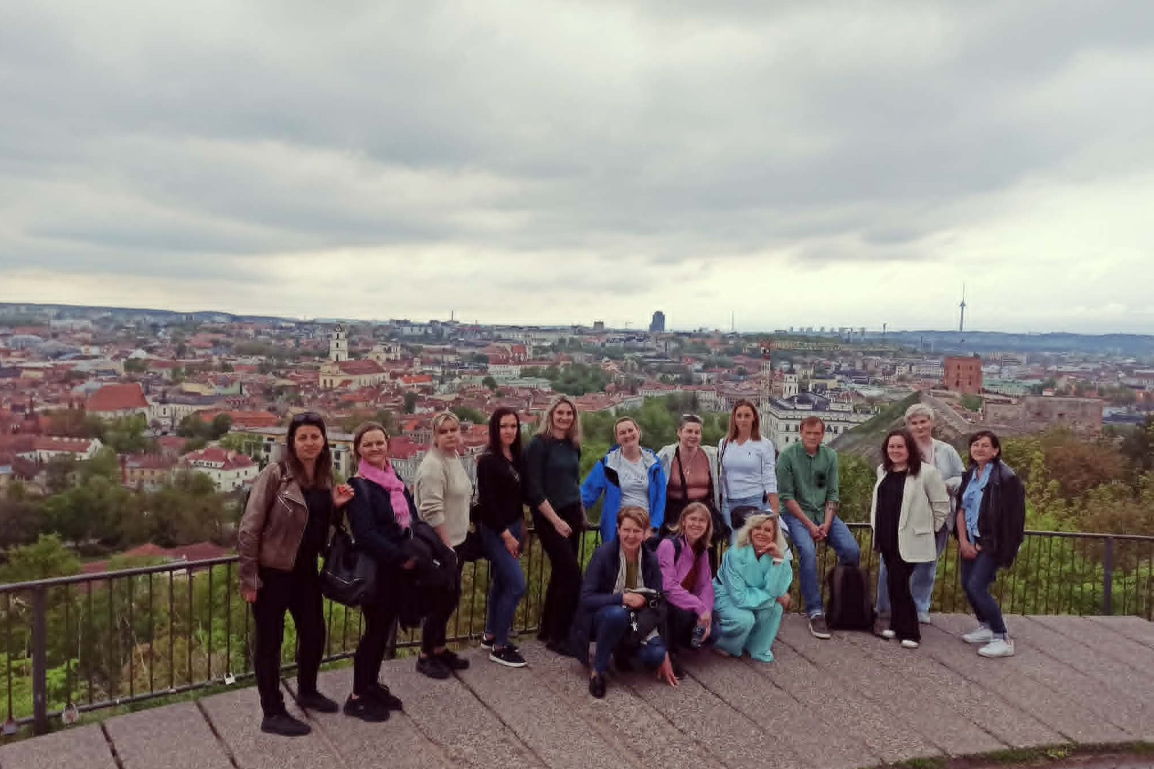 Grupė žmonių pozuoja apžvalgos aikštelėje ant Trijų Kryžių kalno. Už jų matyti Vilniaus senamiesčio panorama, Gedimino kalnas su pilies bokštu ir debesuotas dangus.