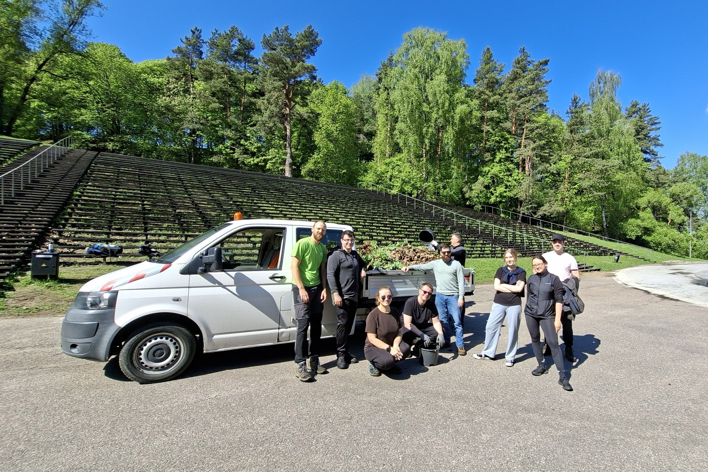 Grupė žmonių stovi lauke šalia balto automobilio su priekaba, kurioje sukrauti lapai ir šakos, o už jų matyti didelės lauko tribūnos ir žalias miškas po giedru mėlynu dangumi.
