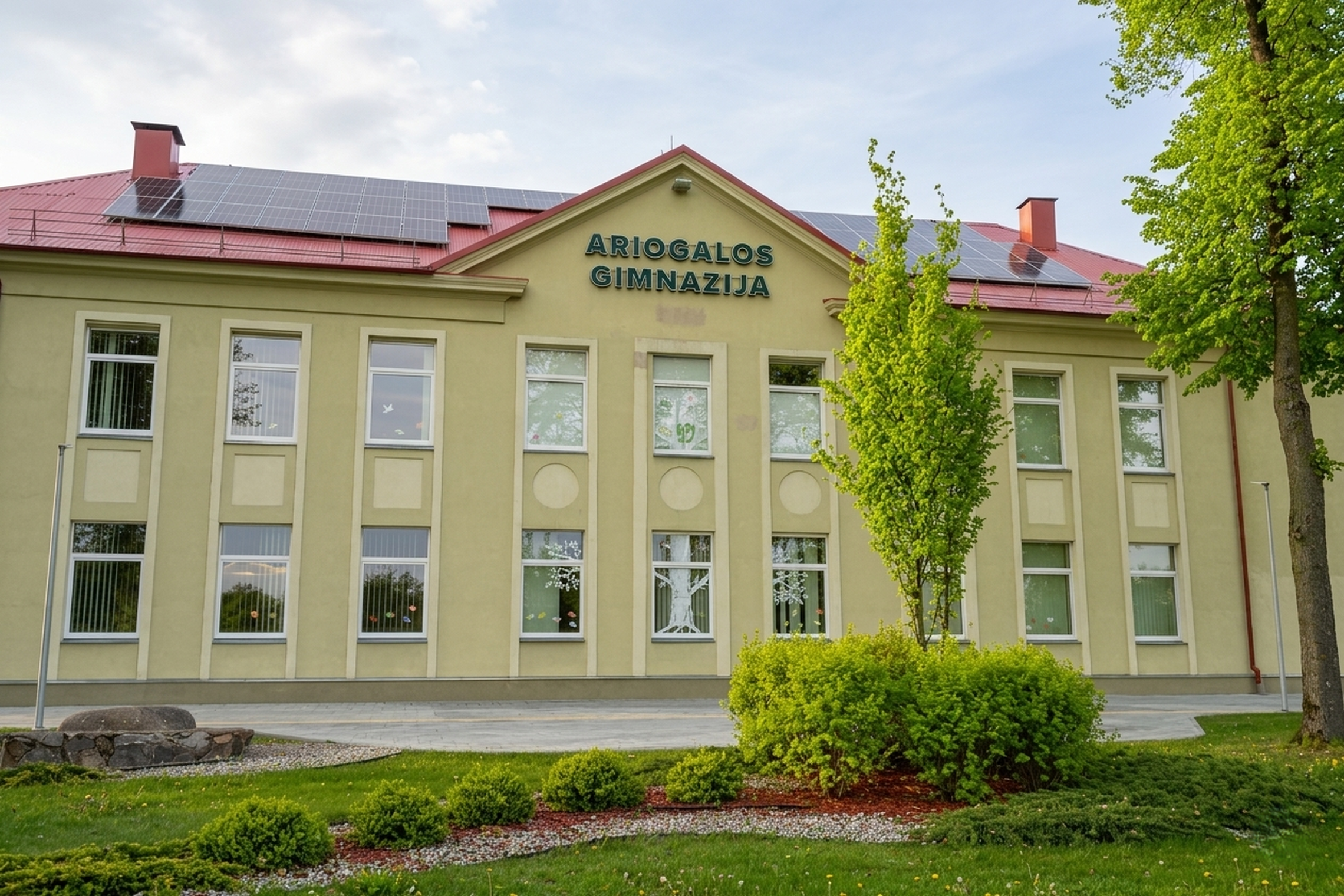 Ariogalos gimnazijos pastatas su užrašu ant fasado, raudonu stogu ir saulės baterijomis. Priekyje matyti sutvarkyta žalia aplinka, medžiai, krūmai ir takas.