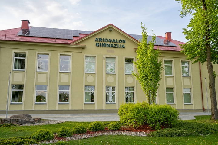 Ariogalos gimnazijos pastatas su užrašu ant fasado, raudonu stogu ir saulės baterijomis. Priekyje matyti sutvarkyta žalia aplinka, medžiai, krūmai ir takas.