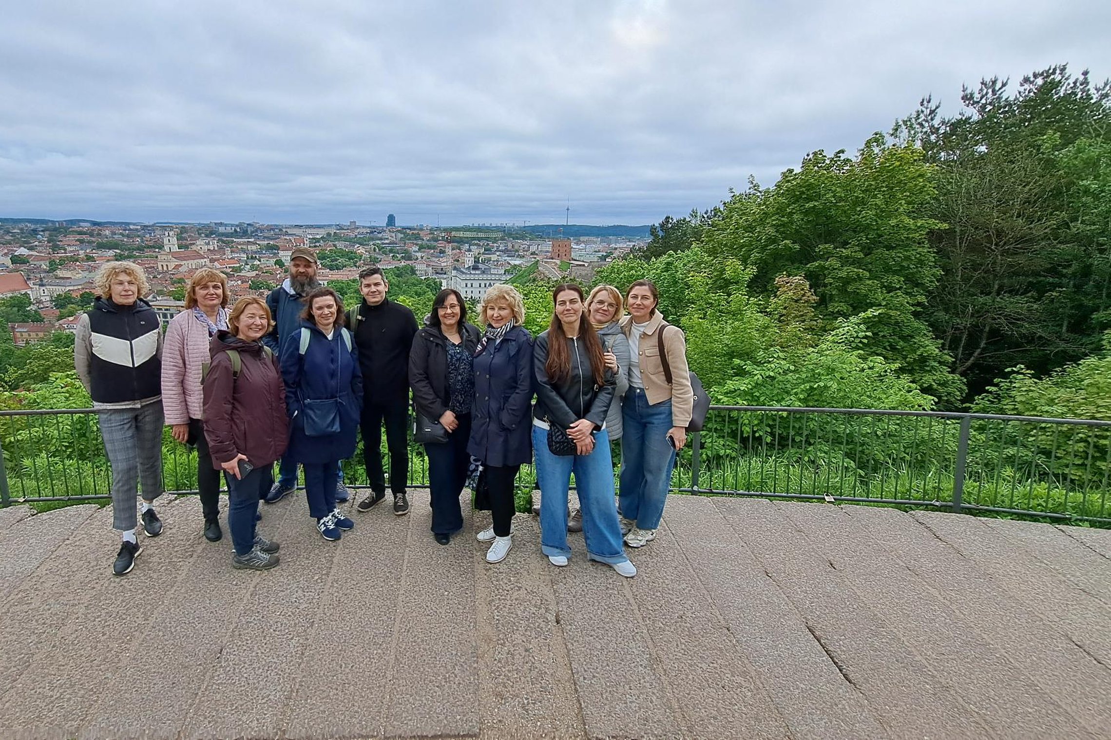 Ekskursijos dalyviai stovi apžvalgos aikštelėje Vilniaus pilių teritorijoje, o fone atsiveria Vilniaus senamiesčio panorama ir žaliuojantys miesto šlaitai. Grupė fotografuojasi po ekskursijos apie Gediminaičių sostų kovas.
