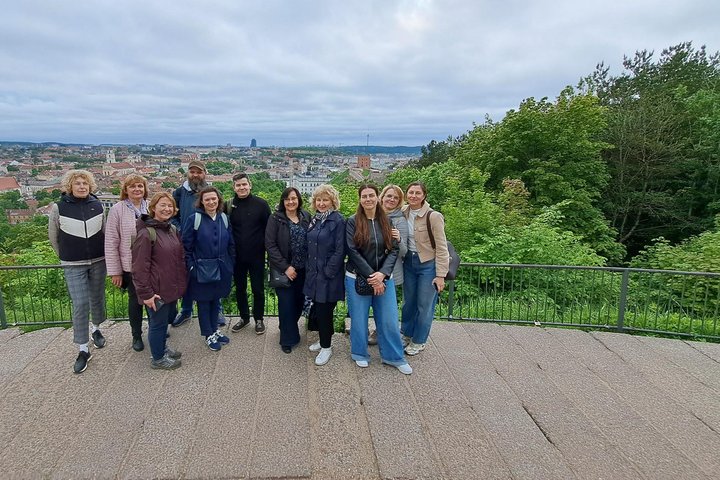 Ekskursijos dalyviai stovi apžvalgos aikštelėje Vilniaus pilių teritorijoje, o fone atsiveria Vilniaus senamiesčio panorama ir žaliuojantys miesto šlaitai. Grupė fotografuojasi po ekskursijos apie Gediminaičių sostų kovas.