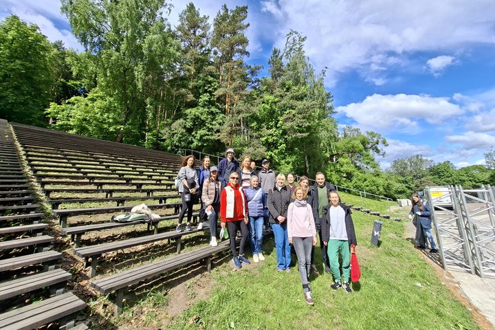 Grupė savanorių stovi lauko amfiteatro tribūnose prie miško, šalia matyti renginio konstrukcijos ir įrangos elementai saulėtą dieną.