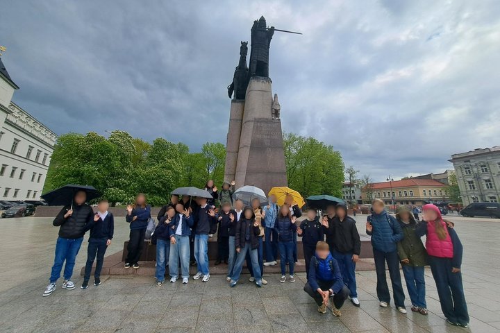 Mokiniai su skėčiais stovi prie kunigaikščio Gedimino paminklo Katedros aikštėje – edukacija apie Vilniaus tris pilis vyko lietingą pavasario dieną. Fone žydi kažtonai.