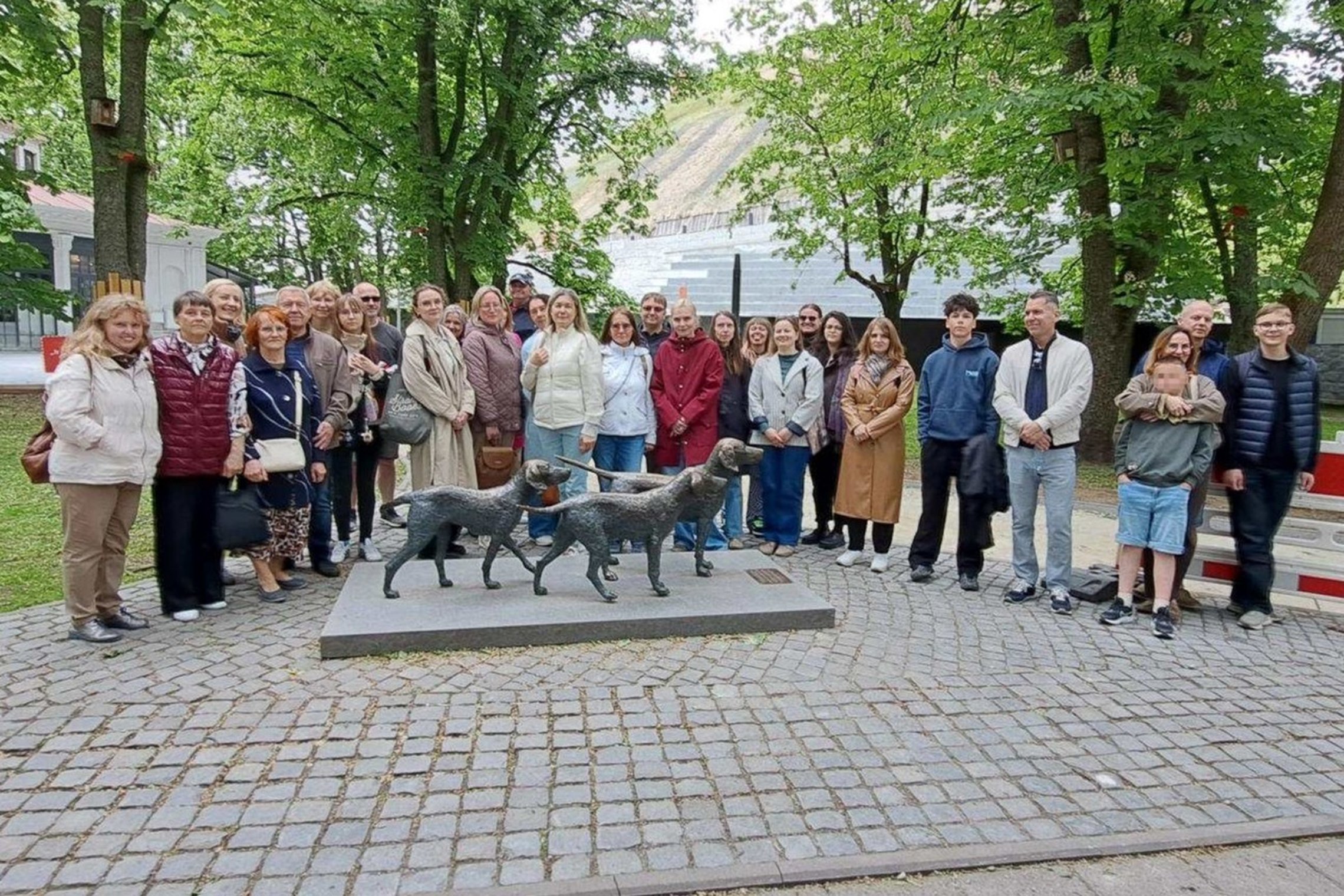 Ekskursijos „Valdovas spaudžia leteną: Vilniaus pilių šunys“ dalyviai pozuoja bendrai nuotraukai prie šunų skulptūros. Žaliuojančių medžių apsuptyje susirinkę dalyviai stovi puslankiu, o priešais juos matyti trijų skalikų skulptūra.