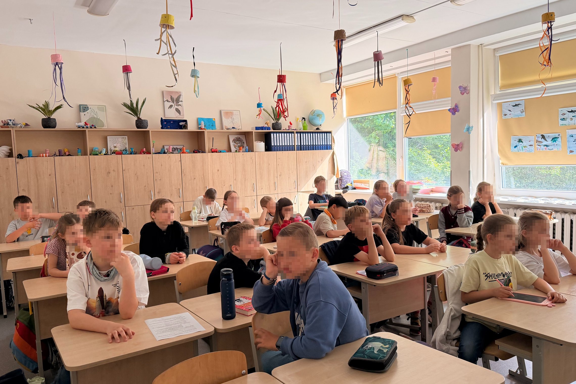 Alt tekstas: Pradinių klasių mokiniai Sereikiškių parke tarp žydinčių medžių klausosi edukacijos apie Lietuvos simbolius ir didžiuosius kunigaikščius, laikydami savo užrašus ir stebėdami pavasarinę gamtą.