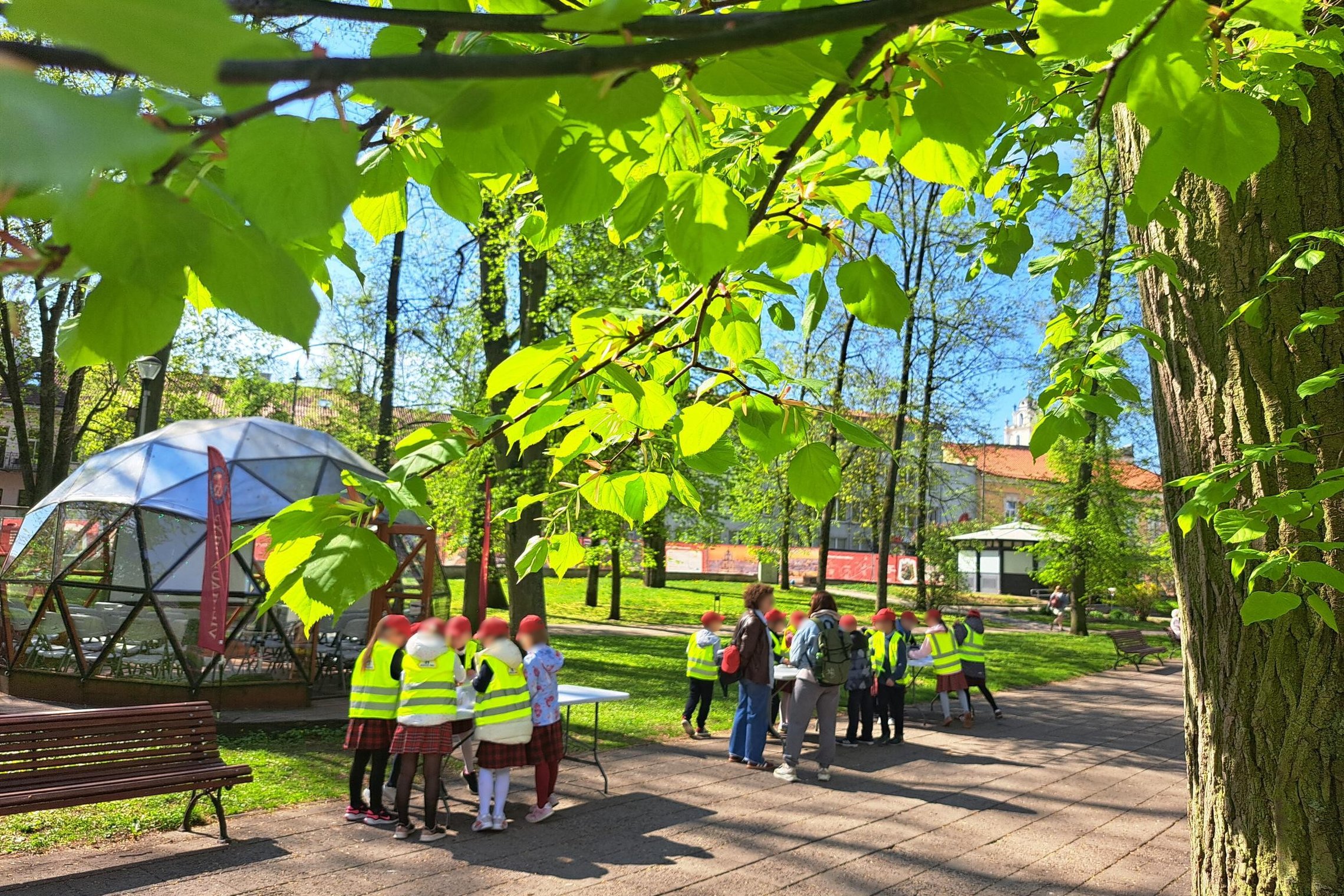Vaikai su ryškiomis liemenėmis dalyvauja edukacijoje šalia Vilniaus pilių kupolo – dirba grupėse prie stalų, o virš jų žaliuoja pavasariniai medžiai.