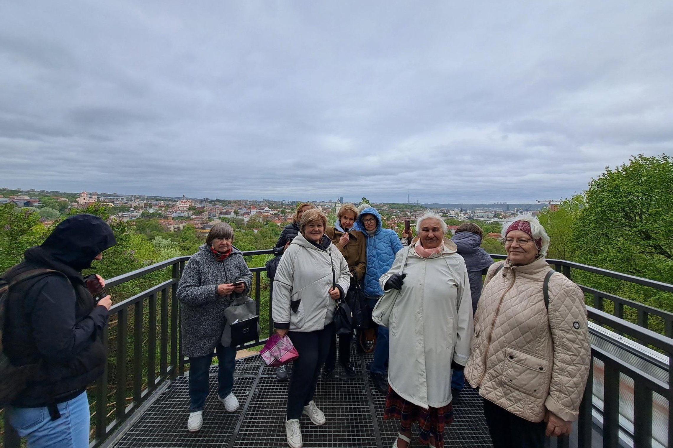 Ekskursijos dalyviai stovi Altanos bokštelio terasoje, už jų atsiveria debesuoto dangaus ir Vilniaus panorama.