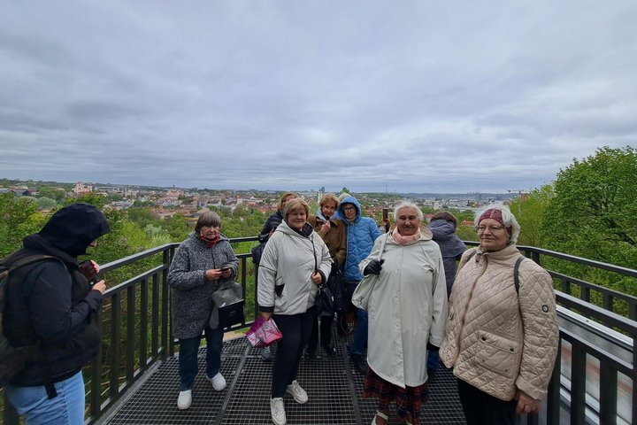 Ekskursijos dalyviai stovi Altanos bokštelio terasoje, už jų atsiveria debesuoto dangaus ir Vilniaus panorama.