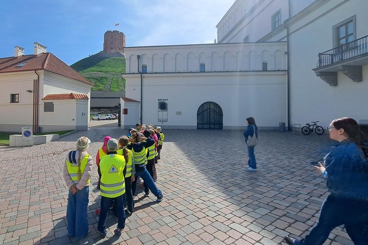 Vaikų grupė su ryškiomis liemenėmis klausosi edukacijos Vilniaus pilių teritorija kieme, fone matomas Gedimino pilies bokštas ant kalno.