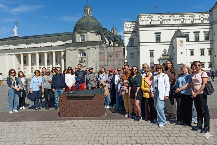 Didelė žmonių grupė pozuoja saulėtą dieną aikštėje priešais monumentą ir istorinius baltus pastatus su kupolu. Priekyje matyti akmenimis grįsta aikštė ir žemas postamentas, o fone – mėlynas dangus.