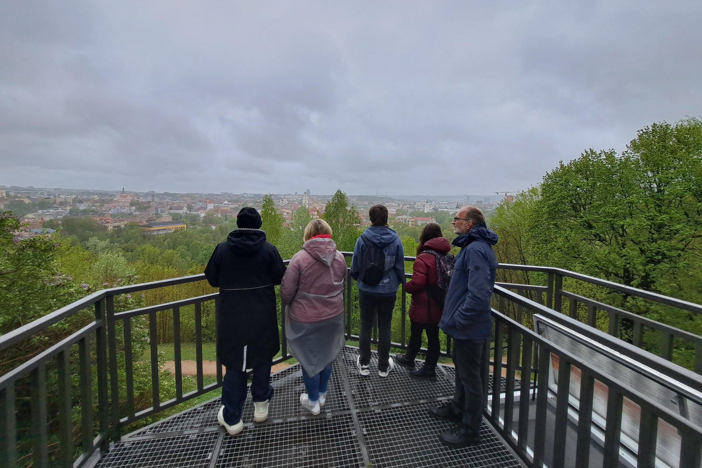 Ekskursijos dalyviai stovi Altanos bokštelio terasoje, už jų atsiveria lietingo ir debesuoto Vilniaus panorama.