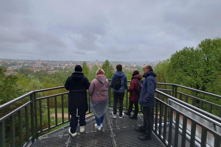 Ekskursijos dalyviai stovi Altanos bokštelio terasoje, už jų atsiveria lietingo ir debesuoto Vilniaus panorama.
