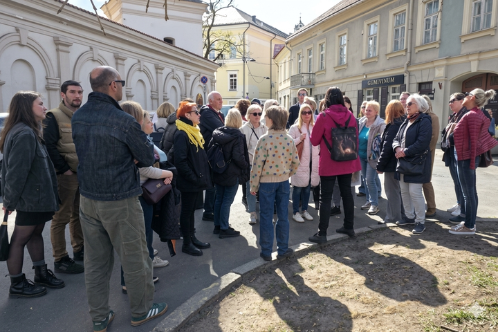 Ekskursijos dalyviai sustoję Vilniaus senamiestyje prie istorinių pastatų. Grupė klausosi pasakojimo, žmonės apsirengę pavasariniais lauko drabužiais, šviečia saulė, ant grindinio ir žemės krinta ilgi šešėliai.